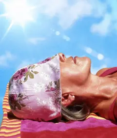 Una mujer recostada tomando el sol con un gorro morado, con flores moradas, que le cubren los ojos.