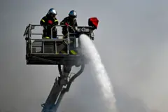 Two firefighters on a platform with a water hose and a ladder with rescue basket.