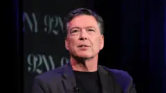 Джеймс Коми в черном пиджаке и черной водолазке с небольшим микрофоном на лацкане.