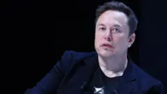 Elon Musk arimo kuvuga areba muri 'camera', ari mu iserukiramuco ry'i Cannes mu Bufaransa, mu mwaka wa 2024. Yambaye ikoti ry'umukara, imbere harimo umupira.