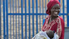 سيدة سودانية نازحة تحمل رضيعها وهي تبتسم