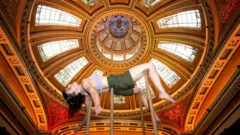Acrobat Alyssa Moore is held up by poles with an ornate ceiling in the background