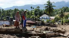 スカーフを頭に巻いた女性3人が、洪水で押し流され泥だらけになった場所に立ち尽くしている