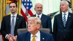 ホワイトうハウスの大統領執務室の机に座るトランプ氏と、その後ろに立つ政府高官ら