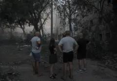 Tres hombres en pantalones cortos y camisetas y una mujer joven con un vestido de verano se encuentran en un patio y miran escombros humeantes después de un ataque en Kramatorsk atribuido al ejército ruso.