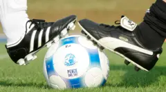 El balón, blanco y azul, con el mapa de Herzogenaurach y los sellos de Adidas y Puma, descanzando sobre una corona de laurel, y la frase Paz, un día. Sobre él, un pie con zapatillas de Adidas y otro con zapatillas de Puma, ambas blanco y negro pero con franjas distintas, todo sobre césped. Al fondo, árboles y cielo azul con nubes. 
