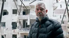 رجل في منتصف العمر يُدعى محمد يقف وسط أنقاض مبنى سكني مدمّر.
