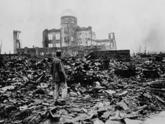 Um homem de costas, entre as ruínas de Hiroshima, diante do esqueleto de um grande edifício à sua frente, depois que uma bomba atômica foi lançada