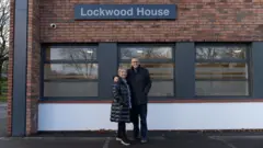 Fotografia de Rob e Dianne Parsons em frente ao Lockwood Centre. Ronnie está à direita de Dianne e usa calças, óculos e um casaco preto. Dianne usa um casaco preto acolchoado e leggings pretas e sorri para a câmera. Uma placa com os dizeres “lockwood house” pode ser vista na frente do prédio de tijolos, acima de uma fileira de janelas.