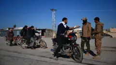 Kırsalda bir caddede üç motorsiklet arka arakaya duruyor, öndekinde bir genç adam var, güvenlik görevlileriyle konuşuyor. İki erkek güvenlik görevlisinden birinin gözleri hariç yüzü kapalı ve şapka giyiyor. Arkadaki motorsiklerde sırayla iki ve bir kişi var bekliyorlar. 