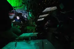 O interior da espaçonave Orion aparece sob luz verde, com tripulantes flutuando em microgravidade. Um deles, Christina Koch, usa um laptop, enquanto outro observa painéis de controle, em um ambiente compacto e cheio de equipamentos.