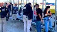 Several law enforcement agents stand near two people whose hands are bound behind their backs. 