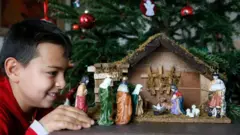 Хлопчик розглядає маленьку шопку зблизька, позаду стоїть прикрашена ялинка