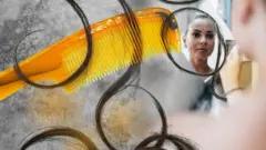 Жінка майже без волосся дивиться в дзеркало, збоку великий гребінець із кучерями