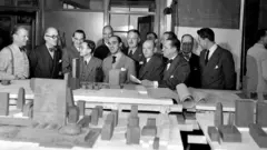 Os membros do conselho responsável pela elaboração dos planos para a construção da sede da ONU em Manhattan. São treze homens de terno, atrás de maquetes do projeto, em uma foto em preto e branco