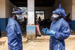 A imagem mostra um prédio parcialmente aberto, com área interna sombreada e chão de terra na parte externa. Em primeiro plano, duas pessoas usam equipamentos completos de proteção, como aventais azuis, luvas, máscaras e viseiras transparentes. Uma delas gesticula com as mãos enquanto a outra a observa, sugerindo uma conversa ou coordenação de trabalho. Ao fundo, várias pessoas aparecem sentadas ou em pé sob a cobertura do prédio, próximas a motocicletas estacionadas e a uma parede com janela