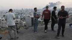 Varios hombres observan desde una colina el humo que sale de la emisora estatal iraní tras ser bombardeada por Israel.