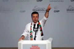 Ekrem İmamoğlu bir kürsüden konuşuyor. Bir elini havaya kaldırmış. Beyaz bir gömleği var. 