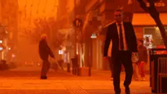 Човек у оделу са маском за лице држи папирну кесу у руци, све око њега је наранџасто, у даљини се назире старији мушкарац