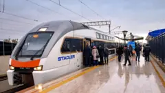 Azərbaycanın Almaniyadan aldığı Stadler markalı yeni elektrik qatarı stansiyada dayanıb və sərnişinlər yağışlı havada qatara minməyə çalışırlar.