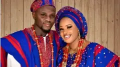 Razak Omotoyossi ati aya rẹ, Bose Alao wọ asọ oke alawọ olomi buluu, wọn fi ilẹkẹ sọrun, ti wọn si de fila ati gele asọ oke naa. Aworan yii ni wọn ya lọjọ Idana fun igbeyawo wọn.