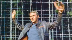 Steve McQueen (1930-1980), aktor AS, mengenakan jaket kulit dan sarung tangan bisbol, dengan kedua tangannya terangkat tanda menyerah, dalam sebuah foto publisitas yang dikeluarkan untuk film, 'The Great Escape', 1963. 