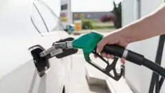 Рука людини тримає паливний пістолет із зеленою ручкою та заправляє білий автомобіль.