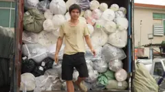 Um homem de camiseta bege e shorts preto está descendo de um caminhão empilhado com rolos de tecido de várias cores - rosa, branco, verde.