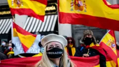 امرأة ترتدي كمامة كُتب عليها "تحرير" وأعلام إسبانية ترفرف خلال مسيرة احتجاجية إلى مبنى مجلس النواب، احتجاجاً على الحكومة ومطالبةً باستقالة الرئيس بيدرو سانشيز، وذلك بالتزامن مع الاحتفال الرئيسي بيوم الدستور الإسباني الذي يُقام في مجلس النواب. 