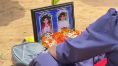2026 මුල්ලිවෛක්කාල් සැමරුමේ ඡායාරුපයක්. වැලි පොළොව මත ගැහැනු ළමුන් දෙදෙනෙකුගේ ඡායාරූපයක් සහ මල් තබා ඇත. කාන්තාවකගේ බව පෙනෙන අතක් දිස්වේ 