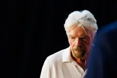 Richard Branson é um homem de cabelos grisalhos. Ele usa uma blusa branca e está olhando para o chão.