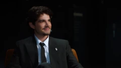 Jão, de cabelos escuros e bigode, veste terno preto, camisa branca e gravata preta, enquanto está sentado em uma poltrona. Ele sorri discretamente e olha para fora do enquadramento, em um ambiente escuro iluminado por luz direcionada.