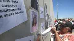 Pessoas participam de protesto em frente à Procuradoria Geral do Estado em Ciudad Juárez, no México, em 2 de agosto de 2025. Os corpos dos seus familiares foram encontrados empilhados em um crematório local, na fronteira norte do México. Eles protestaram para existir justiça e transparência nas investigações. A imagem mostra uma mulher de camisa florida, colando um cartaz em um muro.