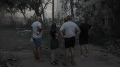 Três homens de shorts e camiseta e uma jovem com roupas leves, de pé em um jardim, observando a fumaça que sai dos destroços causados por um ataque em Kramatorsk, atribuído ao exército russo.