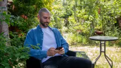 Um homem com bigode e barba escuros com camisa azul, camiseta branca e calça traseira, sentado em um jardim, olhando para o celular.