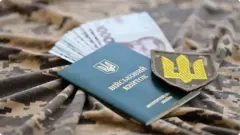 Ілюстративне фото, на піксельній тканині лежить військовий квиток, гривневі купюри, шеврон
