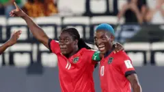 Tabitha Chawinga lève le bras droit en signe de victoire et passe son bras gauche autour de l'épaule de Temwa Chawinga, qui arbore une chevelure bleue et un large sourire. Les deux joueuses portent des maillots rouges du Malawi à liserés noirs : Tabitha avec un brassard de capitaine vert et le numéro 11 blanc sur le devant, et Temwa avec le numéro 10 sur la poitrine. On les voit de face, et une tribune clairsemée se devine en arrière-plan, légèrement floue.