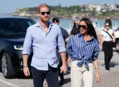 Harry e Megan de mãos dadas durante um passeio. Eles estão usando óculos escuros