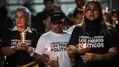 Personas participan en una vigilia para exigir la libertad de los presos políticos venezolanos afuera de la Zona 7 de la prisión de la Policía Nacional Bolivariana (PNB) en Caracas. Se ve a tres mujeres con camisetas blancas y negras, con la inscripción "Liberen a todos los presos políticos" escritosen ellas". Llevan velas en las manos. 