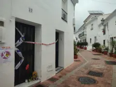 Casa branca em uma rua isolada por fitas policiais; flores são deixadas na porta da frente