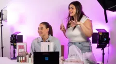 Two young women surrounded by studio lights and tripods selling eyelash serums on a live stream