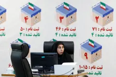 Mulher com trajes típicos em frente a computador; atrás dela, painel mostra ilustração de urnas