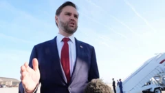 Takım elbiseli bir adam arkada uçağın merdivenleri görünürken gazetecilere konuşuyor. Tek elini kaldırmış