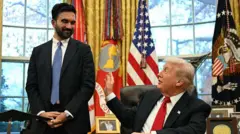 スーツ姿でひげを蓄えたマムダニ氏が、ホワイトハウスの大統領執務室の机に座るトランプ氏の隣に立っている。2人は笑顔で、トランプ氏は右手を伸ばし、冗談めかしてマムダニ氏の左腕を叩いている