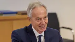Tony Blair yambaye ikositimu y'ubururu na karuvati y'ubururu hamwe n'ishati y'umweru. Afite umusatsi w'imvi. Inyuma ye hari ameza abaje mu giti. 