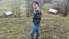 Un hombre en una colina mira a la cámara, vestido con vaqueros y botas, hay graneros y colinas onduladas en la distancia.