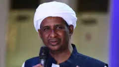 Mohamed Hamdan Dagalo avugira mu ndangururamajwi. Yambaye igitambaro kera n'umwenda ufite ubururu bwijimye ahagana hejuru. 
