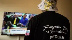 Umutinganyi w'umugore w'Umunya-Uganda, uherutse kwibasirwa ubu akaba acumbikiwe, arimo kureba televiziyo igaragaza ikoraniro ry'inteko ishingamategeko ya Uganda ryiga ku mushinga w'itegeko rirwanya abatinganyi, aha ari mu kigo gifasha aba LGBTQ hafi y'i Kampala, ku itariki ya 21 y'ukwezi kwa gatatu mu 2023. 