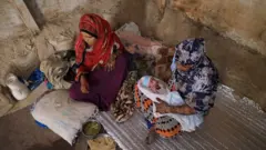 أم صهيب مع رضيعها وامرأة في غرفة بسيطة في مخيم جبل زيد