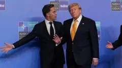 ダークスーツ姿のルッテ氏とトランプ氏が並んで立ち、トランプ氏は右手を上げてカメラを見ている。ルッテ氏は右腕を広げ、左の方を見ている